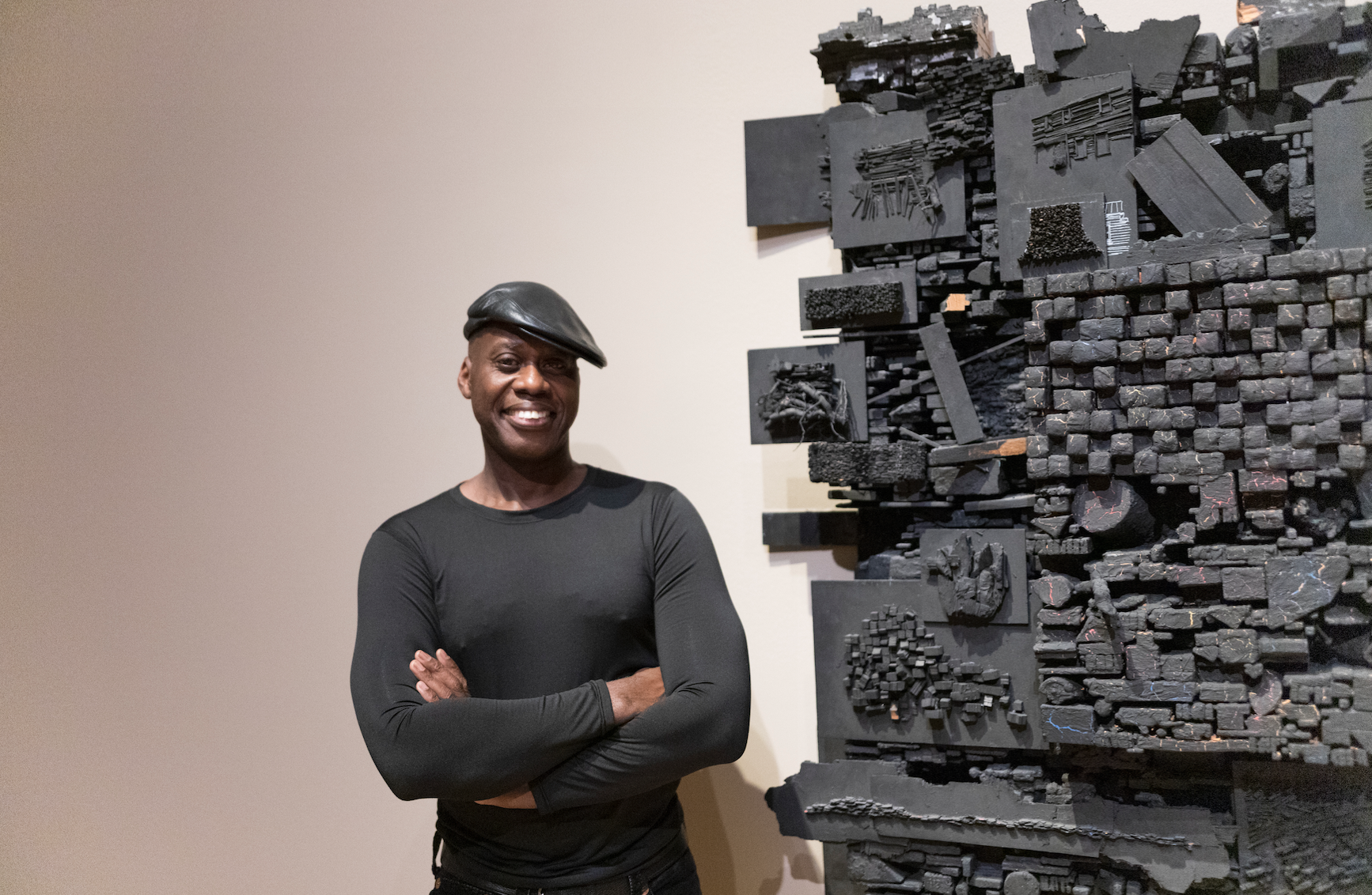 Photograph of artist Leonardo Drew next to his sculpture made of black wood pieces