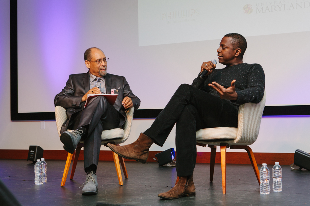 Artist Sanford Biggers and UMD professor Curlee Holton seated on a stage during an event, with Sanford holding a microphone