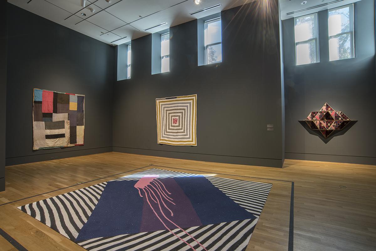 Museum gallery with two large square quilts and one three-dimensional quilt on the walls, with sand-quilt on the floor