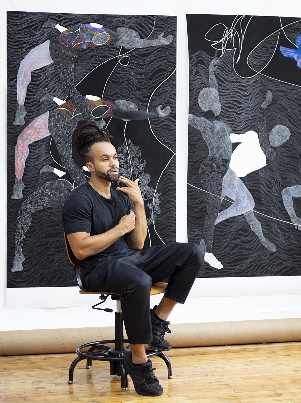 Photo of Nate Lewis sitting on a stool in front of two of his artworks on the wall