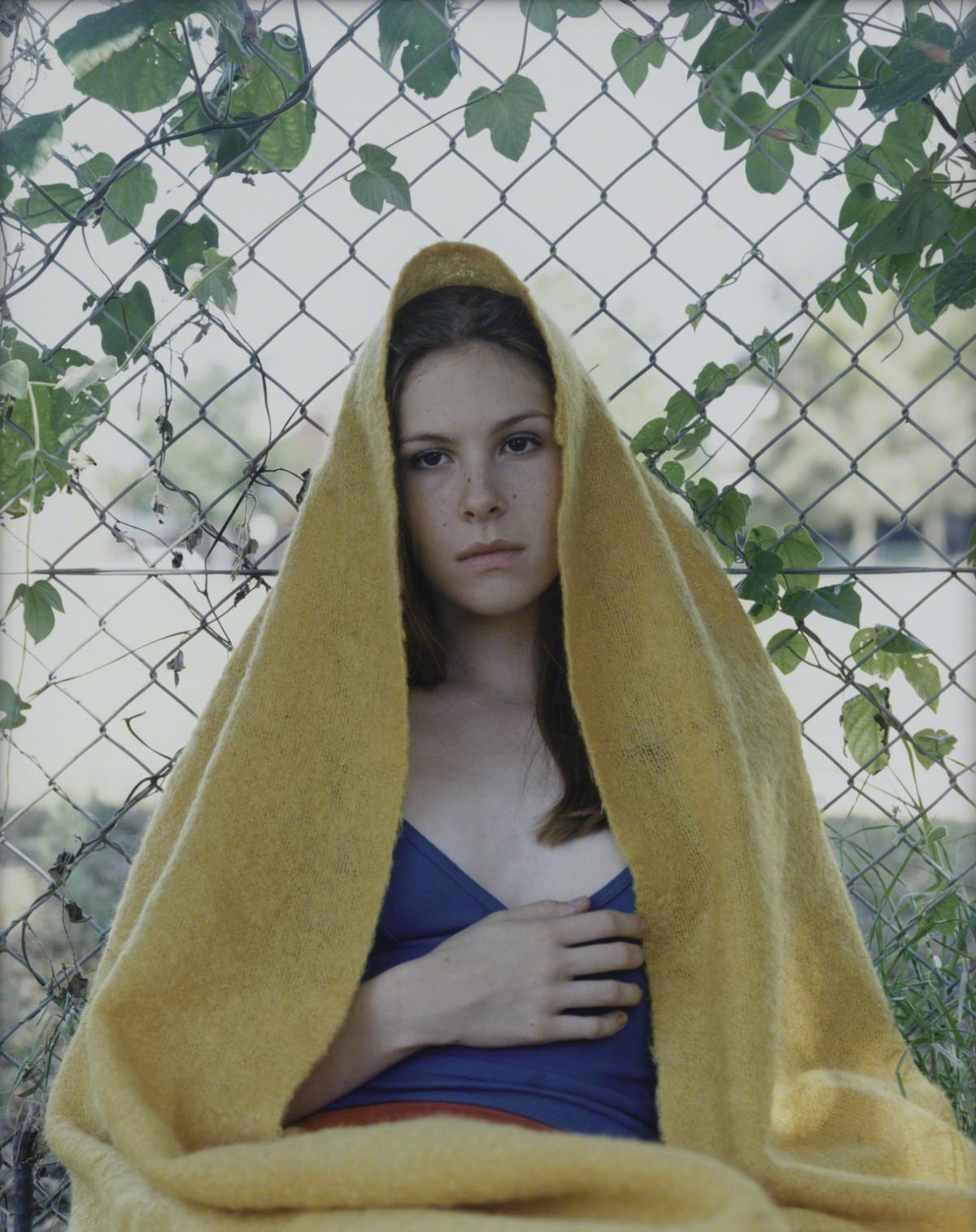 Color photograph of a young woman with a yellow blanket draped over her head sitting by a chain link fence