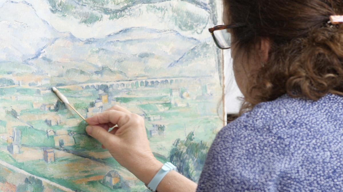 Dark haired woman cleans the surface of a green landscape painting with a cotton swab