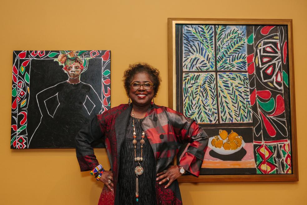 Photograph of a person standing between two paintings, hands on hips and smiling