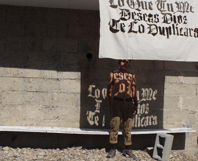 Photograph of Jonathan Herrera Soto standing in front of a building, shirtless, with a cloth with words cut out of it hanging in front of him