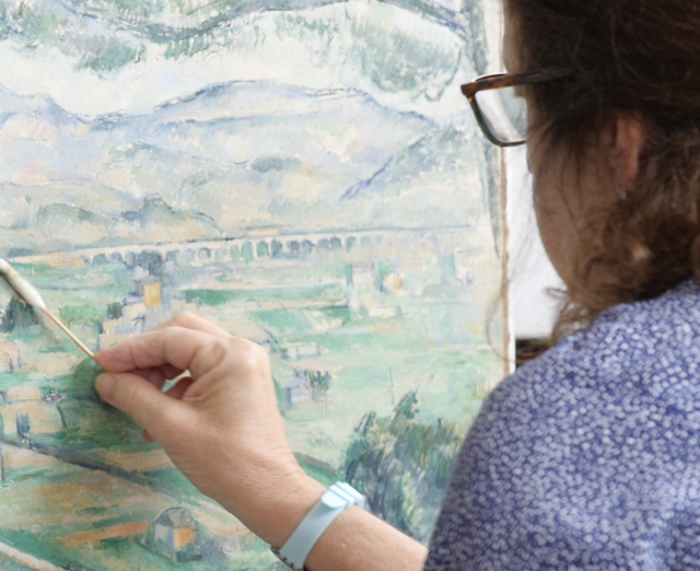 Dark haired woman cleans the surface of a green landscape painting with a cotton swab