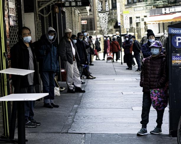 Photograph of people lined up outside on a sidewalk, with masks on