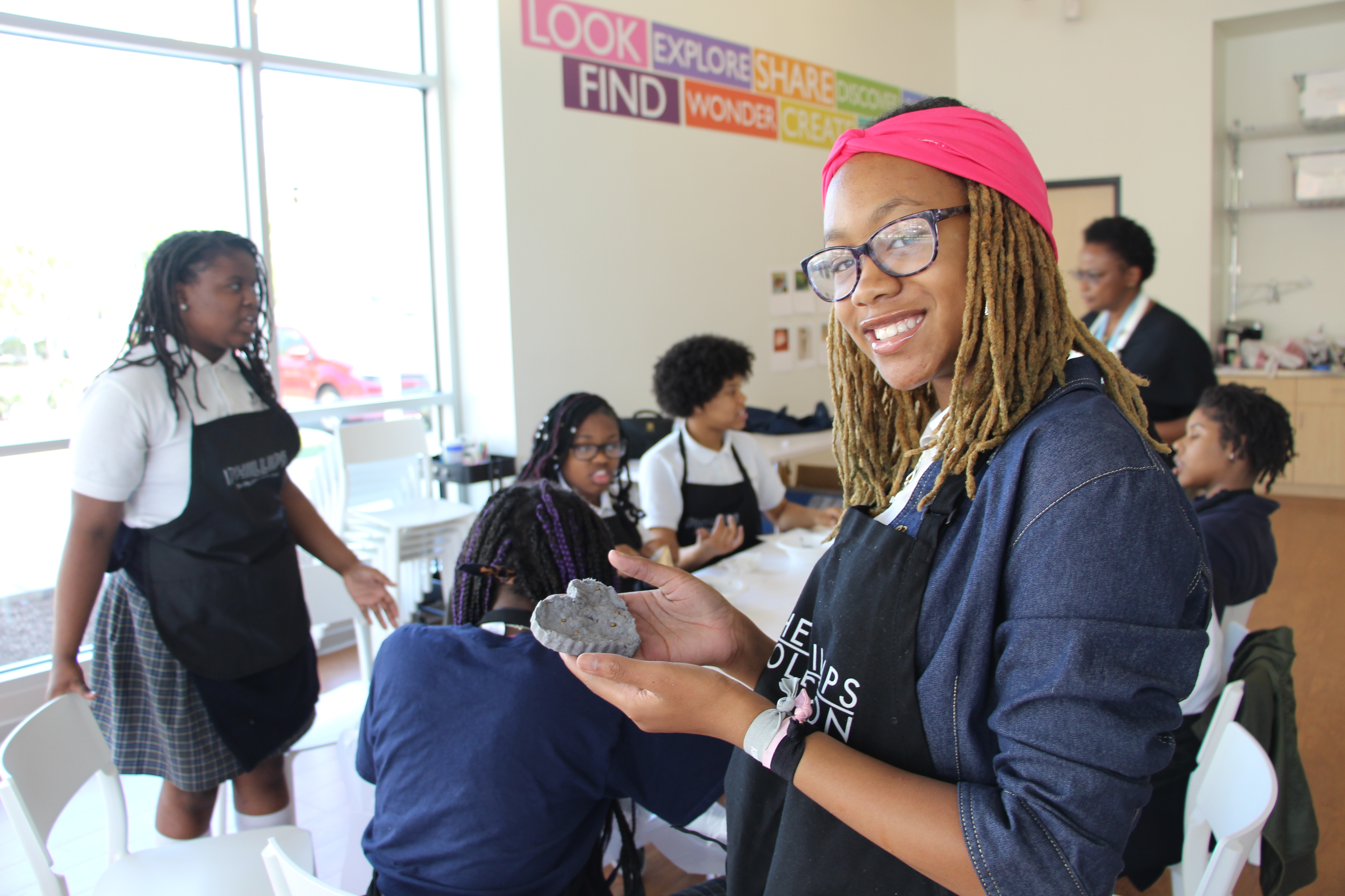 Photograph of female students in an art workshop with one smiling at the camera and holding up her creation