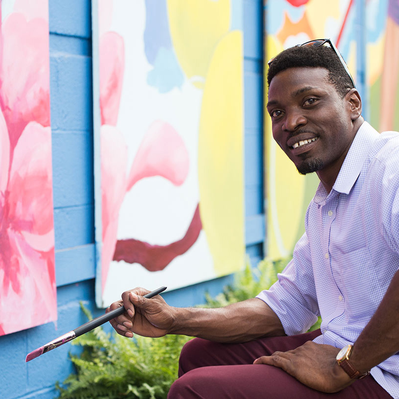 Photo of artist Aniekan Udofia sitting in front of colorful paintings holding a paint brush