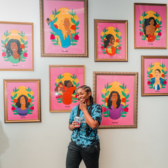 Photo of Trap Bob smiling, standing in front of framed portrait illustrations