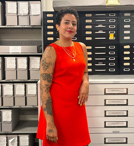 Photograph of Amanda Acosta standing in the archives wearing a red dress