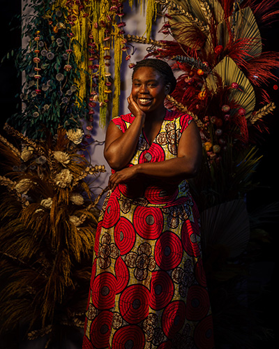 Photograph of Natalie Courtney in a patterned dress with lots of plants and decor behind her