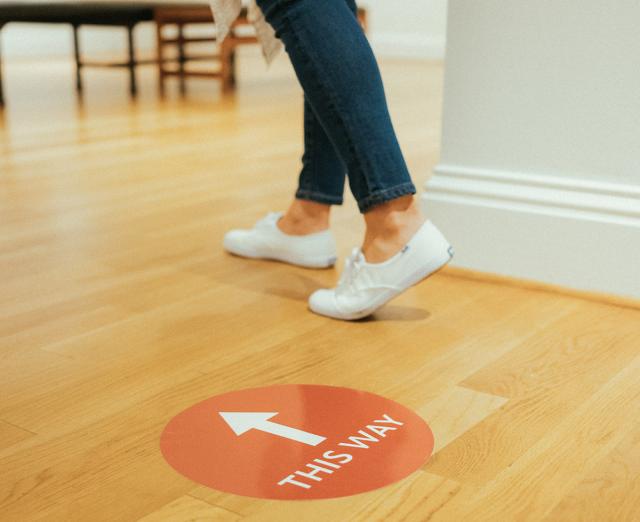 Photograph of a person's legs walking in the direction of a graphic posted on the ground that says "This Way"