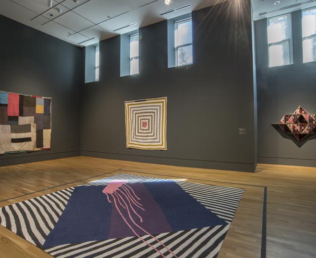 Museum gallery with two large square quilts and one three-dimensional quilt on the walls, with sand-quilt on the floor