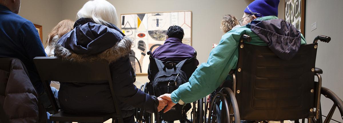 Photograph of two older adults--one in a wheelchair--sitting in a gallery looking at art and holding hands