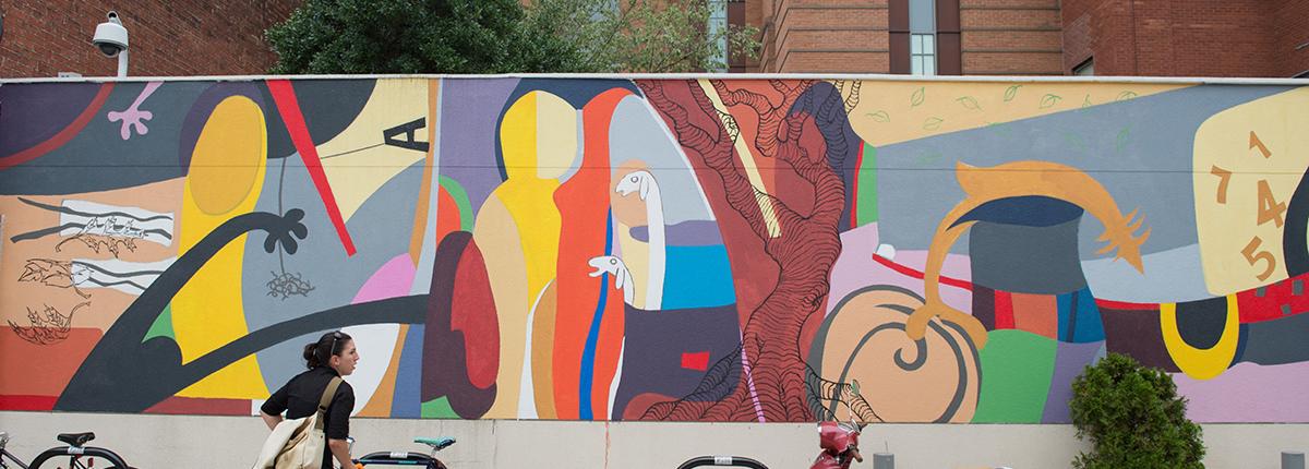 Photograph of a colorful wall mural outdoors, with a woman standing in front by a bike and a motorcycle