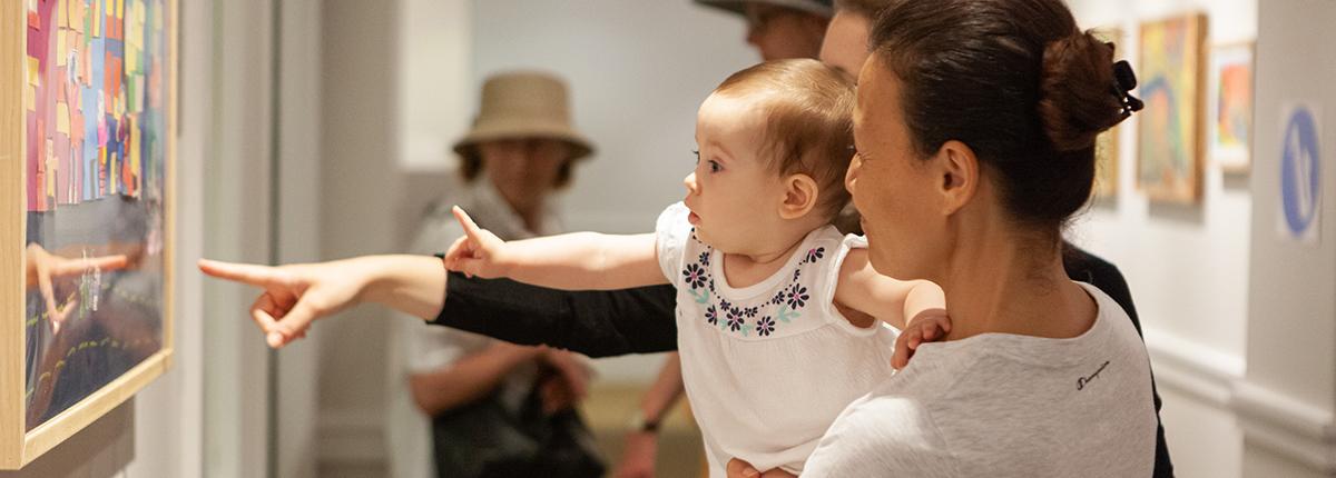 Photograph of woman holding a baby that is pointing at art