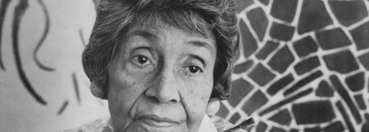 Black and white photograph portrait of Black artist Alma Thomas holding a paint brush with a painting in the background