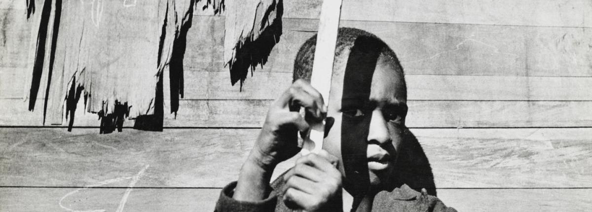 Black and white print of Black boy in a jacket holding a wooden stick in front of a wooden building