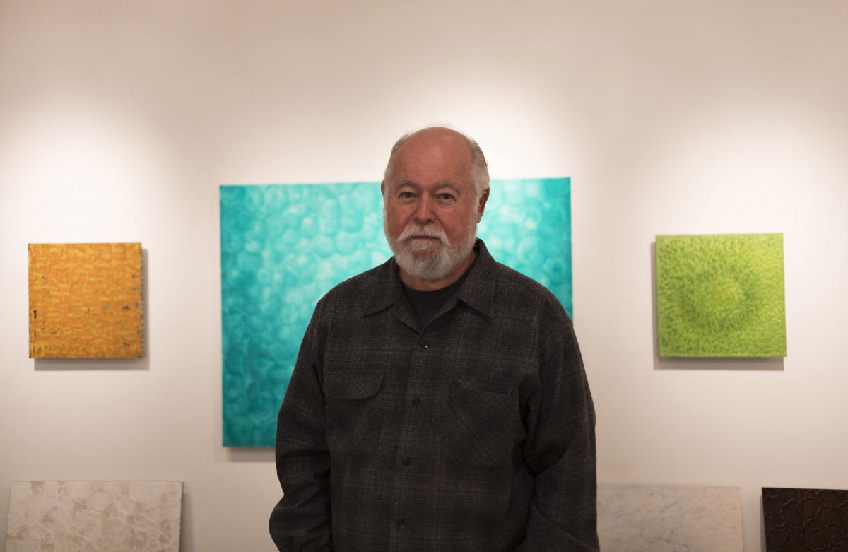 Photograph of Robin Rose with his artwork on the walls behind him