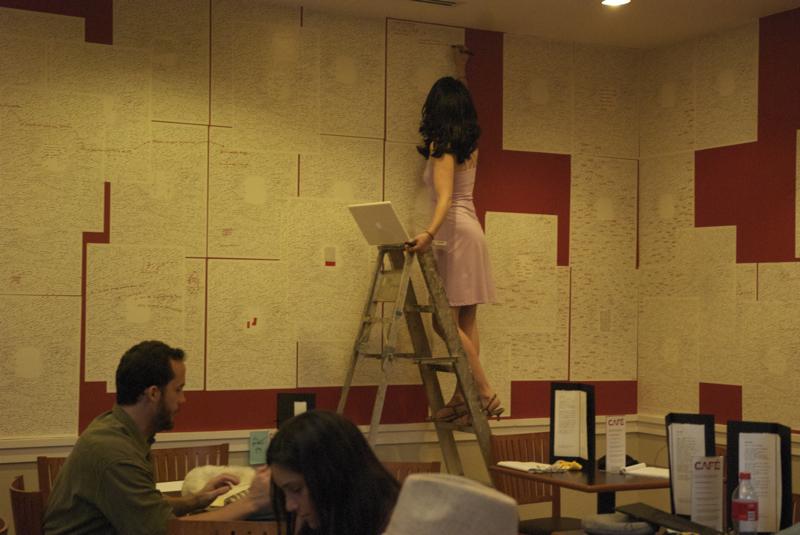 Woman on ladder writes on paper stuck to the wall of a crowded cafe.  