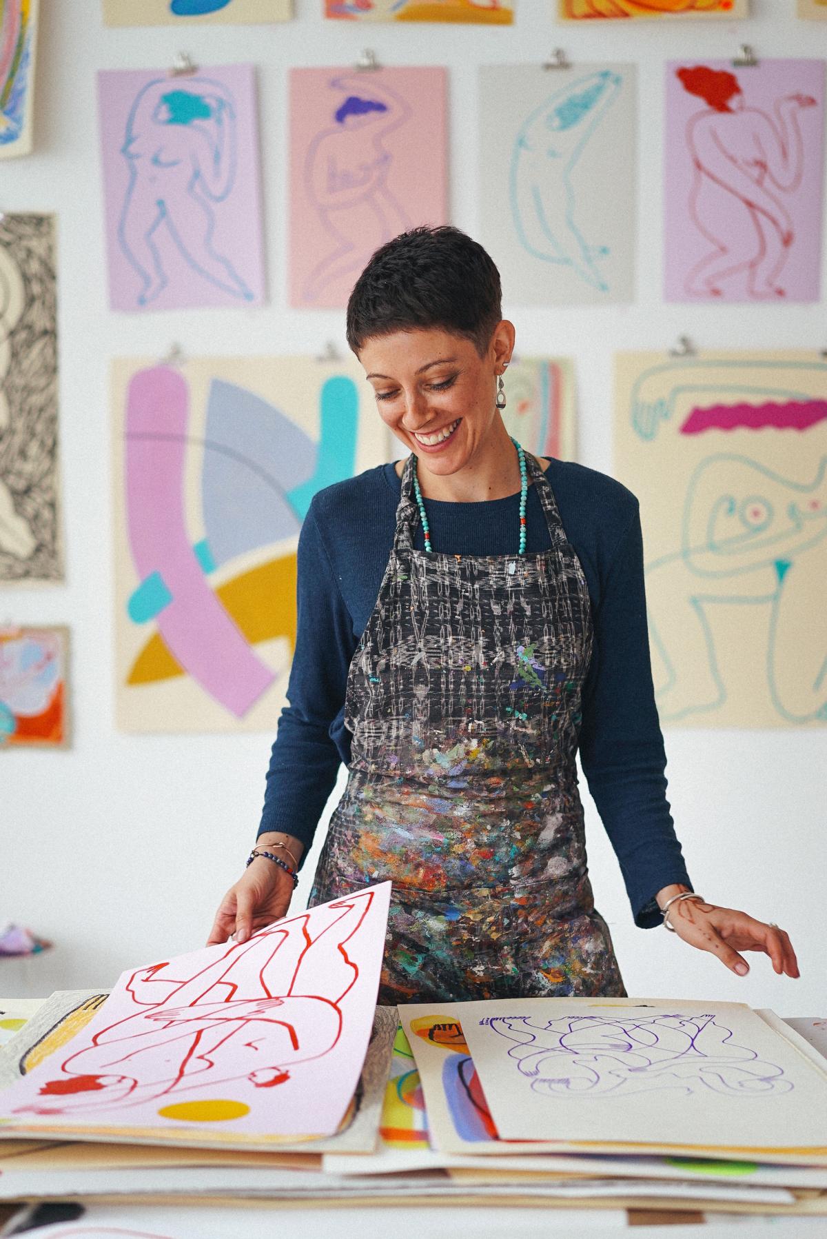 Photograph of a smiling woman standing in a print studio surrounded by colorful prints