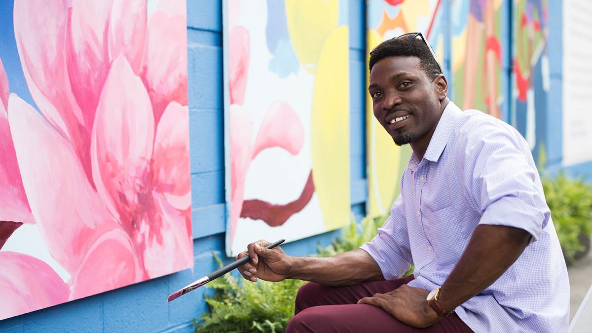 Photo of artist Aniekan Udofia sitting in front of colorful paintings holding a paint brush