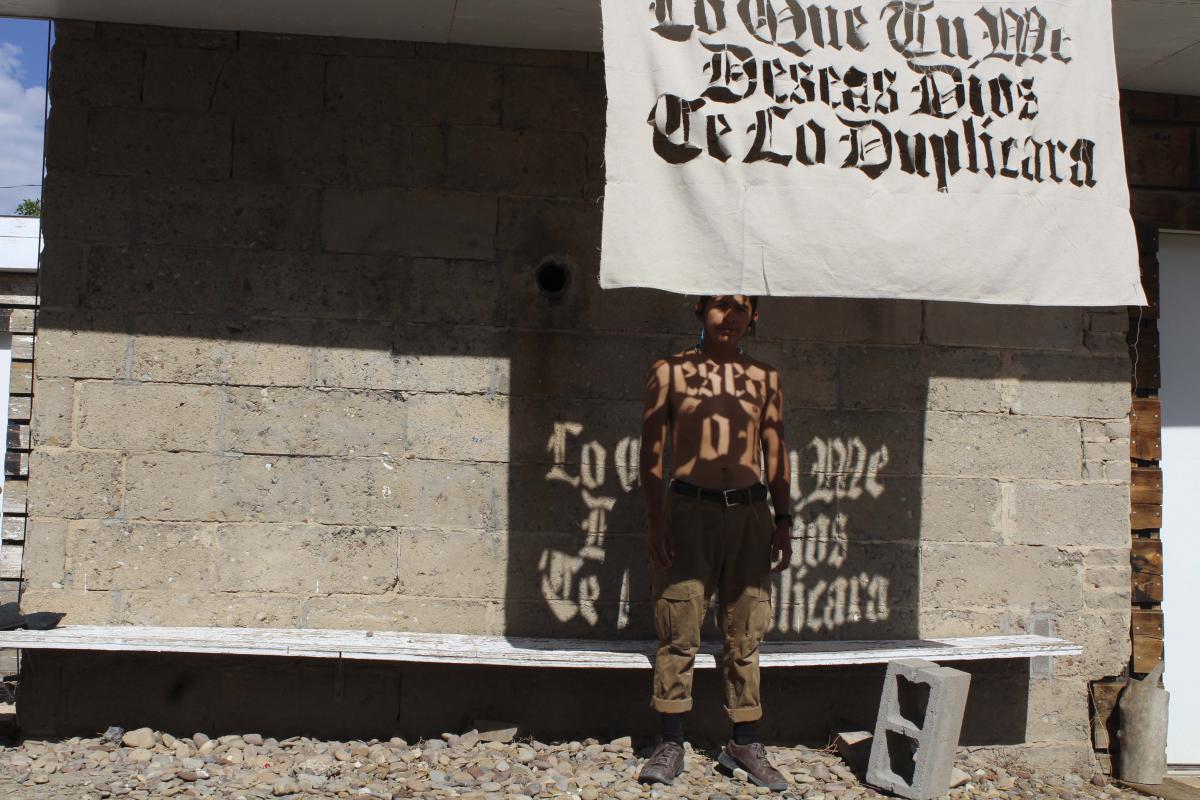 Photograph of Jonathan Herrera Soto standing in front of a building, shirtless, with a cloth with words cut out of it hanging in front of him