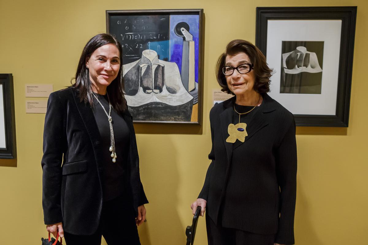Photograph of two women standing with two artworks on the wall, smiling at the camera