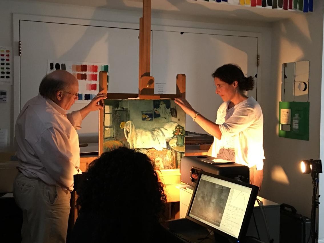 Two people hold a painting on an easel with a light directed on it