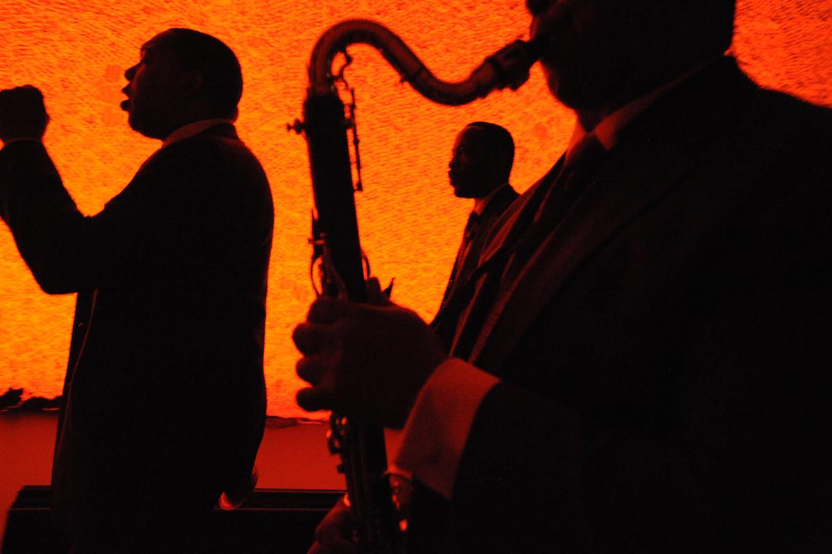 Photograph of three men performing on stage, one with a saxophone, with red orange lighting