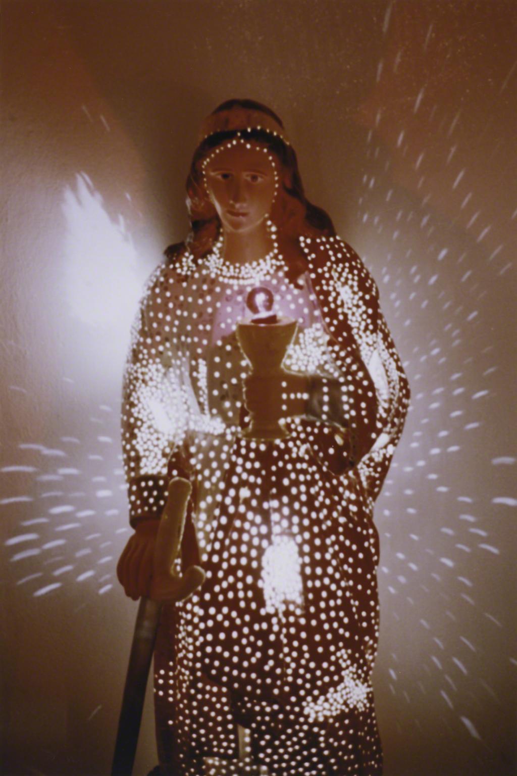 Color photograph of a Christian saint carrying a sword and chalice that is light from the inside and casting light on the wall behind