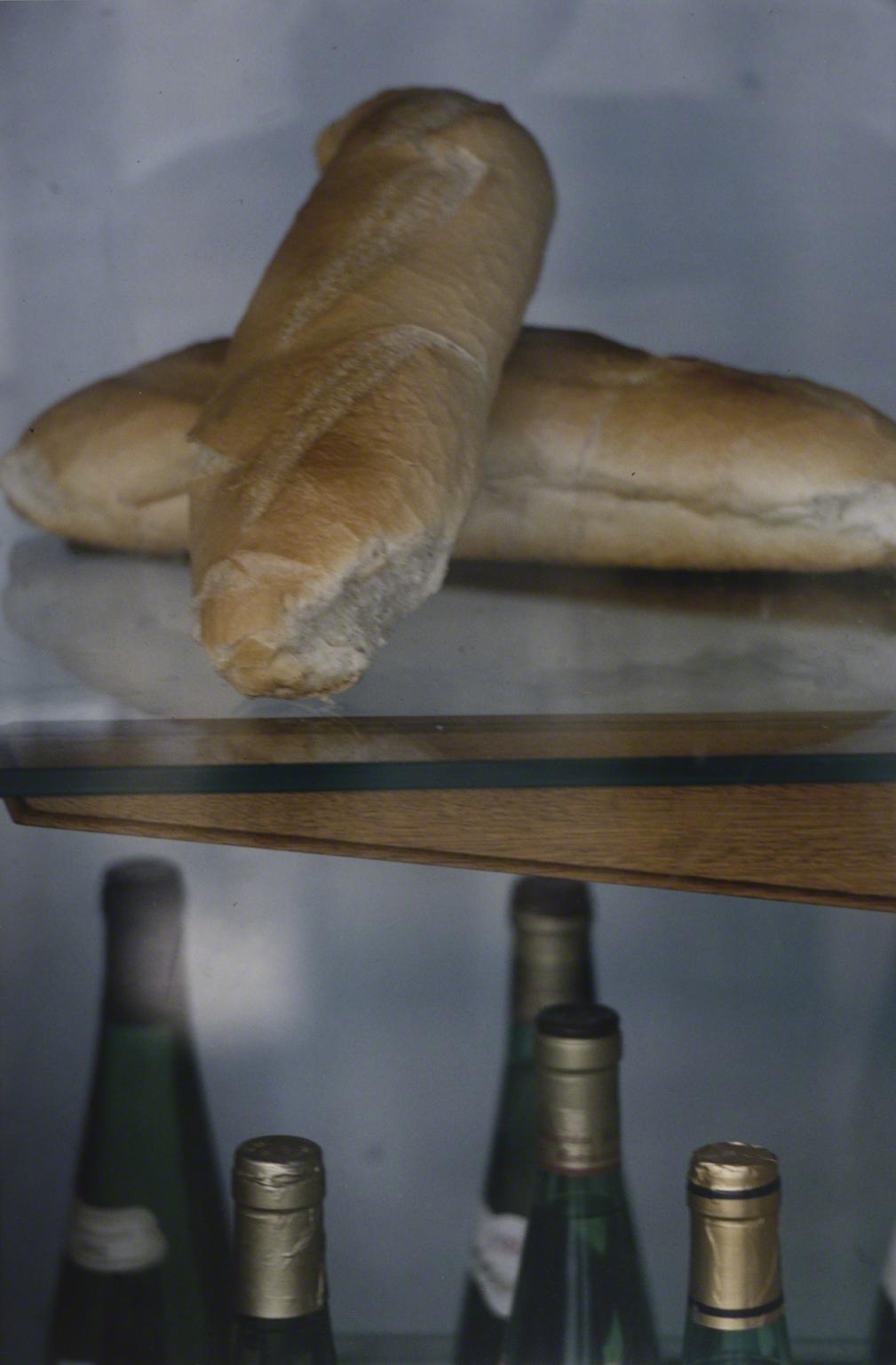 Color photograph of a a glass shelf with two loaves of bread on the top and five bottles on the bottom shelf