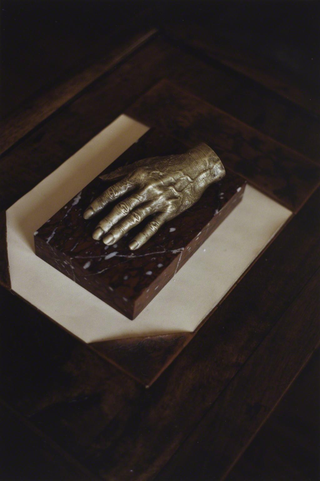 Color photograph of a hand cast in bronze resting on a small slab of red and white marble