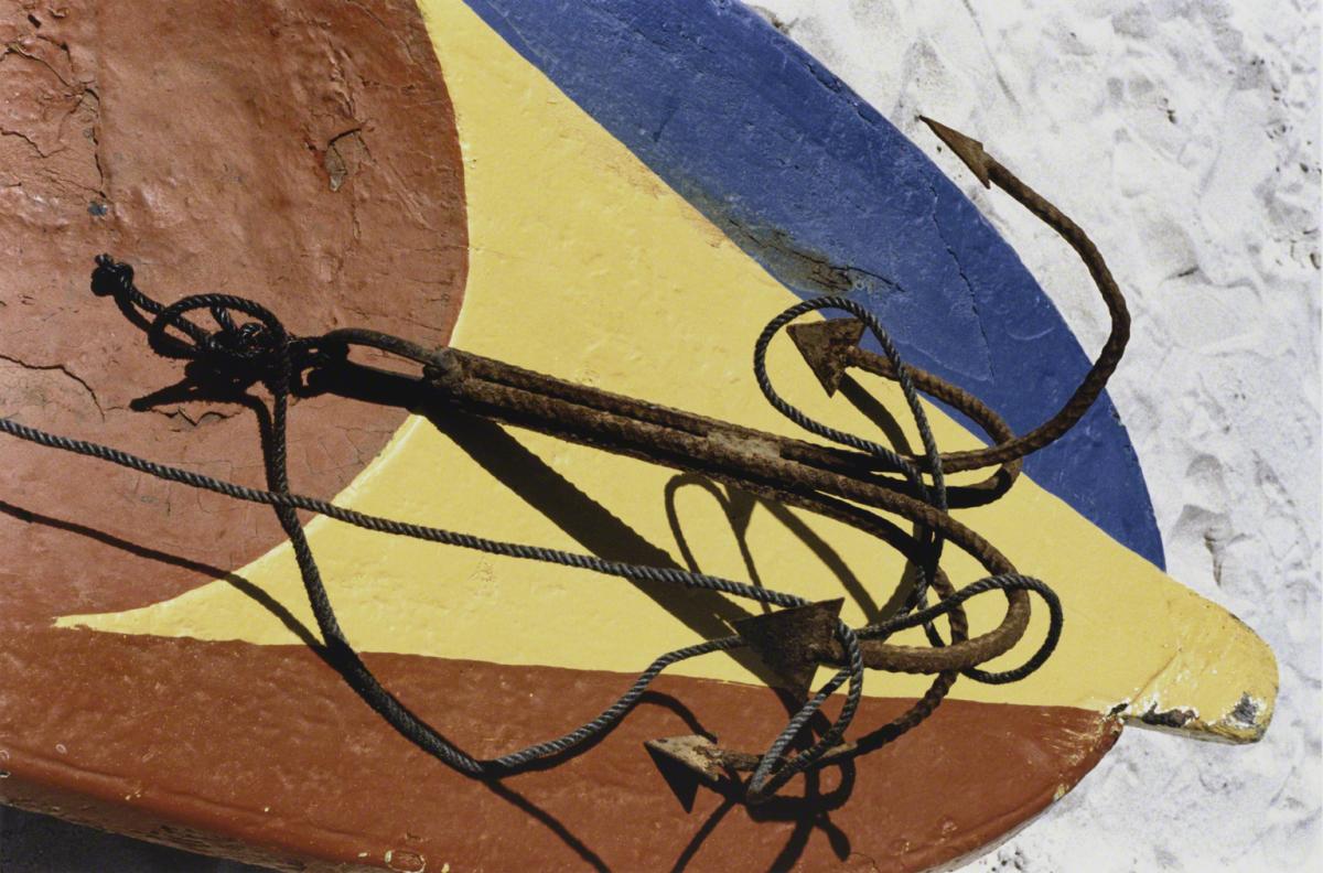 Color photograph of a board painted in blue, red, and yellow with a rusty anchor and background of sand