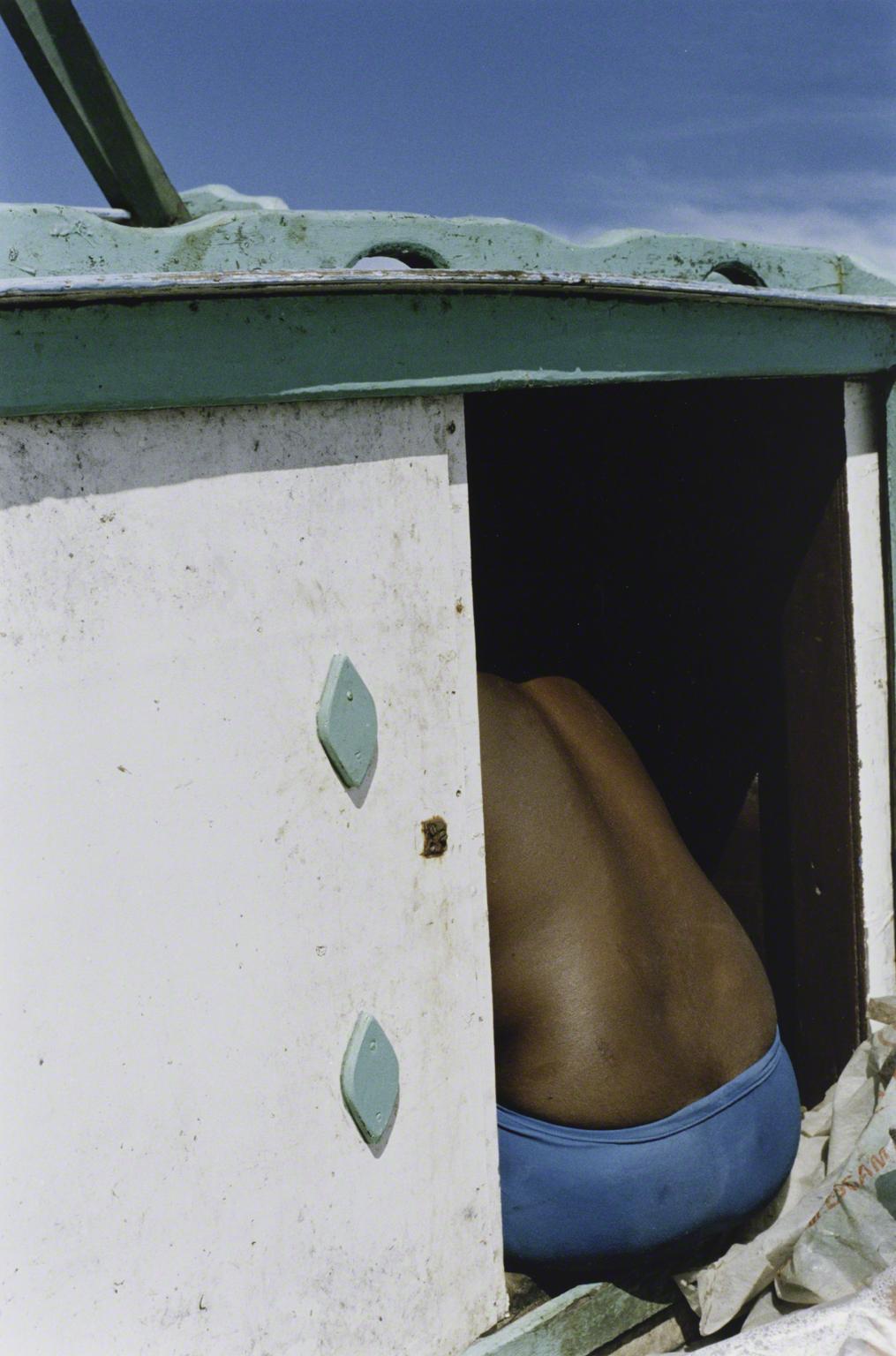 Color photograph of a man with his back to the camera while sitting at the entrance of a boat hatch