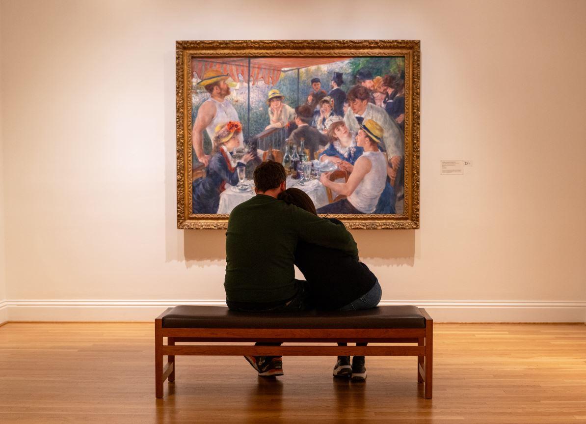 Two people embracing on a bench looking at Renoir's Luncheon of the Boating Party