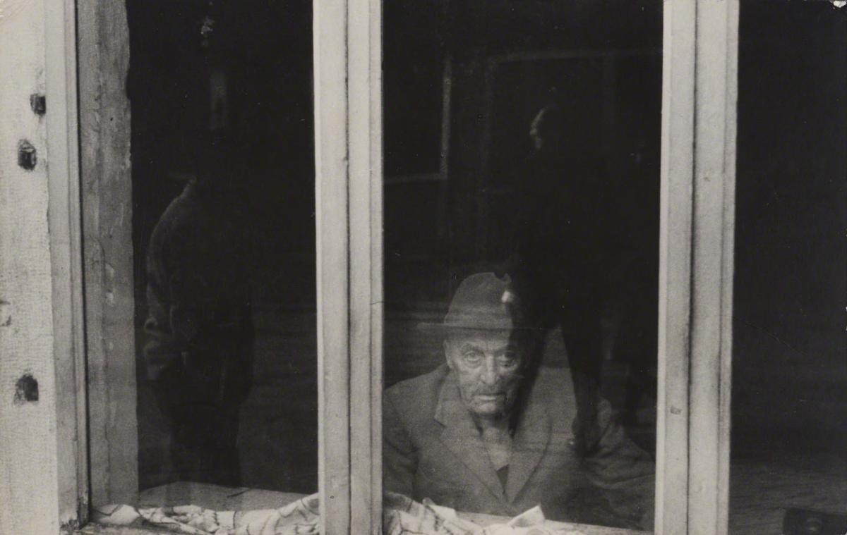 Leon Levinstein, Untitled (man in hat looking through window), 1960.