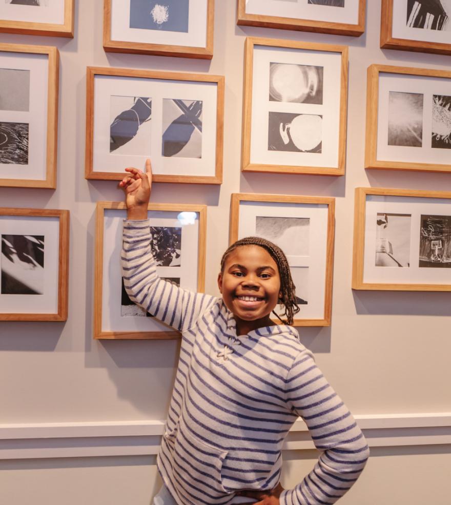 Photograph of a student standing in front of a wall of framed prints, pointing at a print
