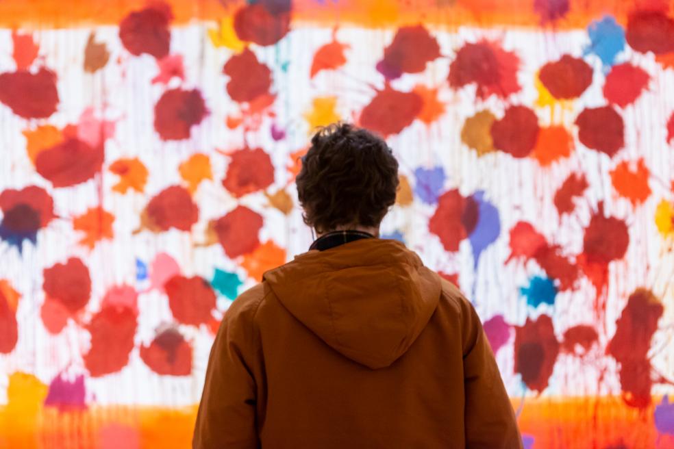 Photograph of a figure looking at a very large artwork with red splotches on a white background