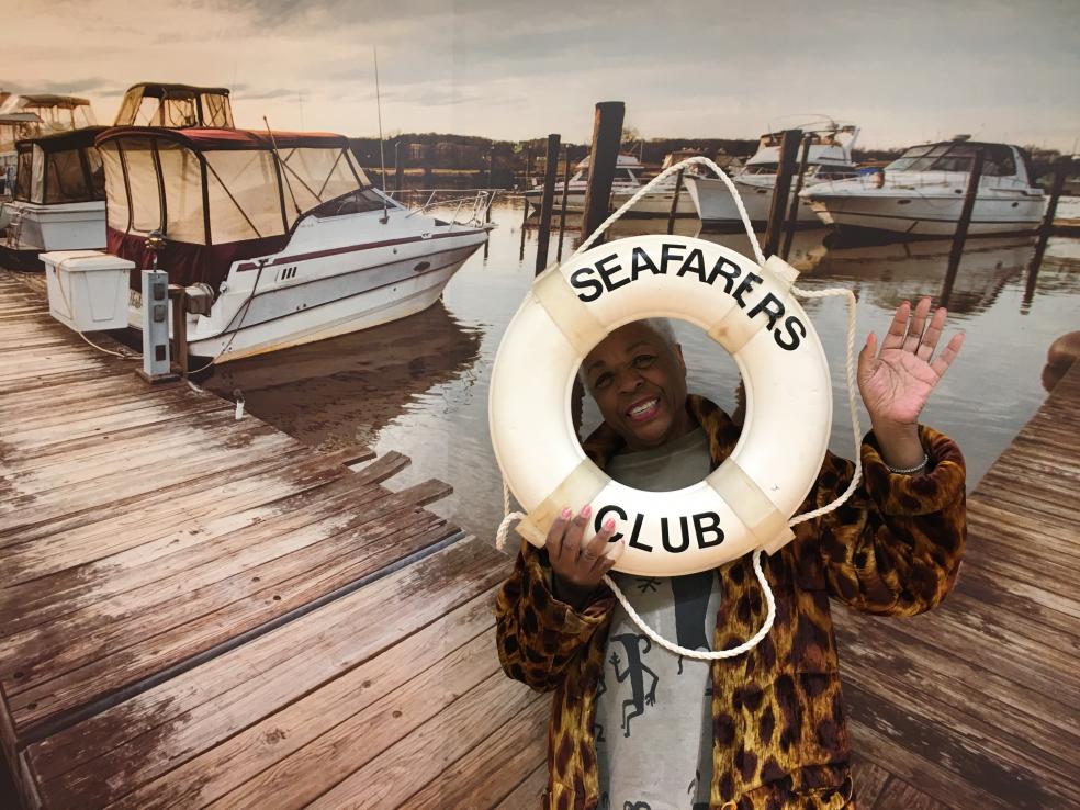 Photo of a woman posing in front of a life ring with the words "Seafarers Club" on it, and standing in front of a printed photo of a dock with boats