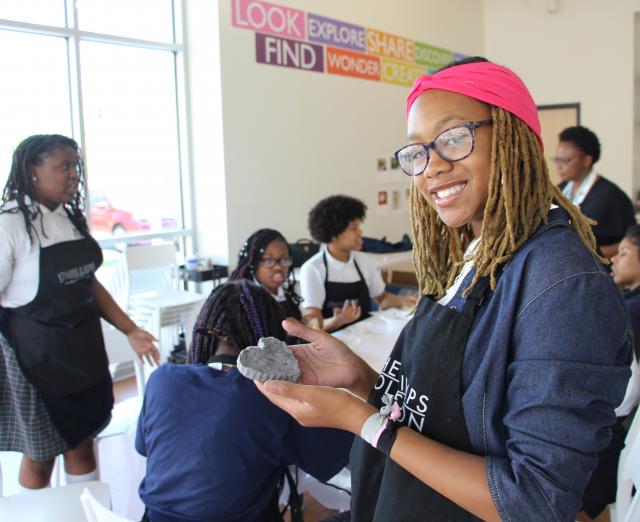 Photograph of female students in an art workshop with one smiling at the camera and holding up her creation