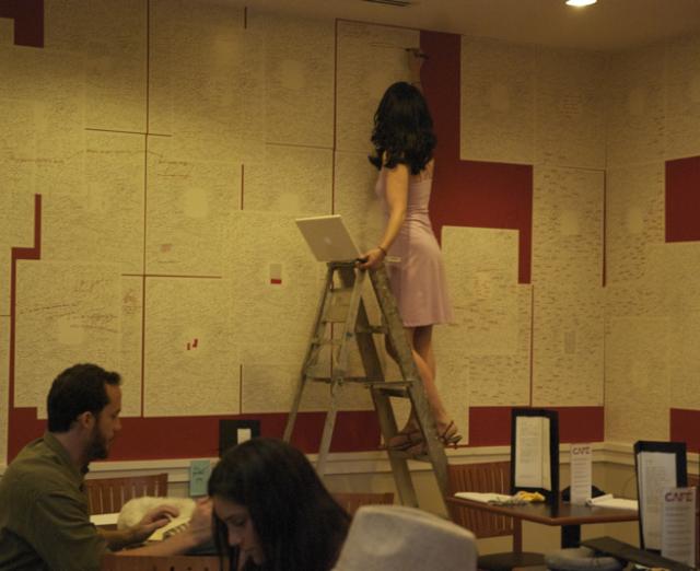 Woman on ladder writes on paper stuck to the wall of a crowded cafe.  