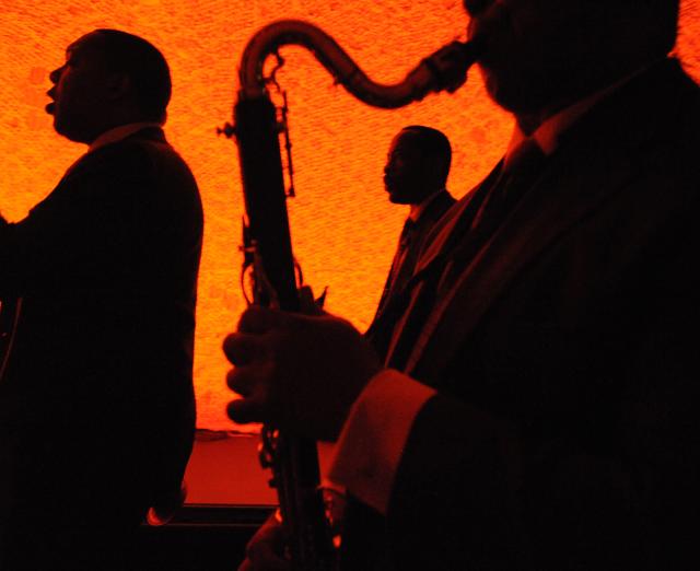Photograph of three men performing on stage, one with a saxophone, with red orange lighting