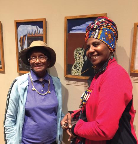 Photograph of two older women smiling in front of panels from Jacob Lawrence's Migration Series.