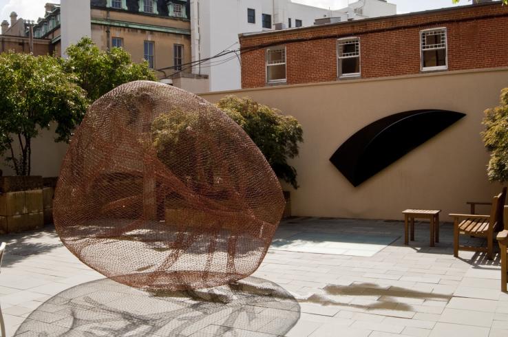Photograph of a large mesh sculpture resting on the ground and a black flat sculpture on a wall in the courtyard