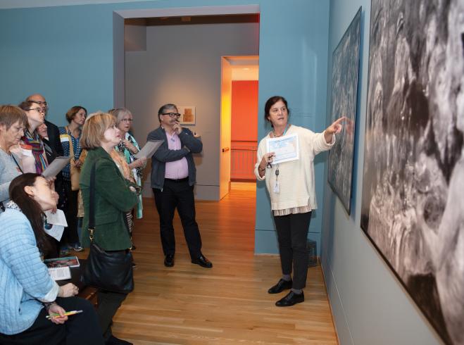 Photograph of a woman standing in front of large print outs of xrays of paintings, speaking in front of a group