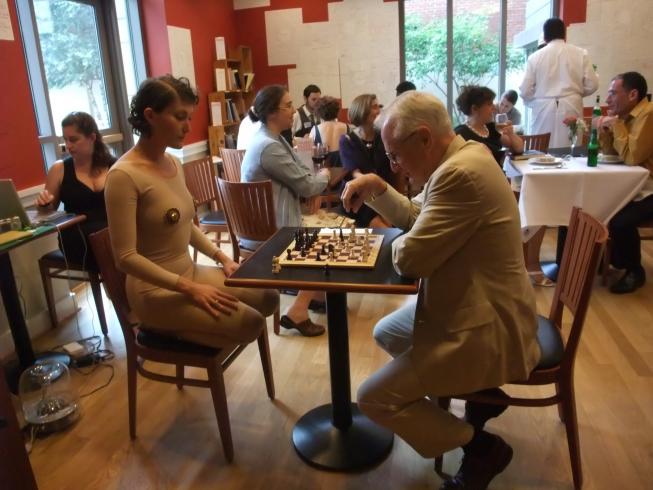 A man and a woman, both dressed in beige, play chess at a cafe table.