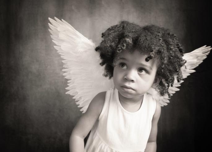 Black and white photograph. A young girl with angelic wings gazes towards the left.