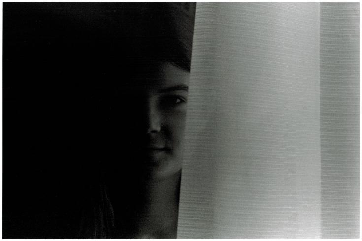 Black and white photograph. A woman's face peers out from dark shadows behind a white curtain.