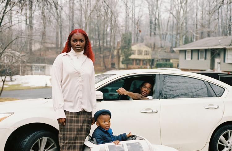 Photograph of a woman and child standing outside a car with a man in the driver's seat